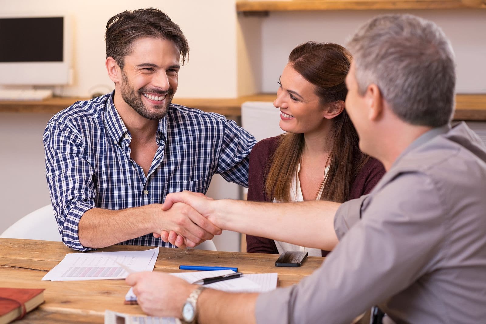 Three people shaking hands across a wooden table during a meeting, with documents and pens visible.