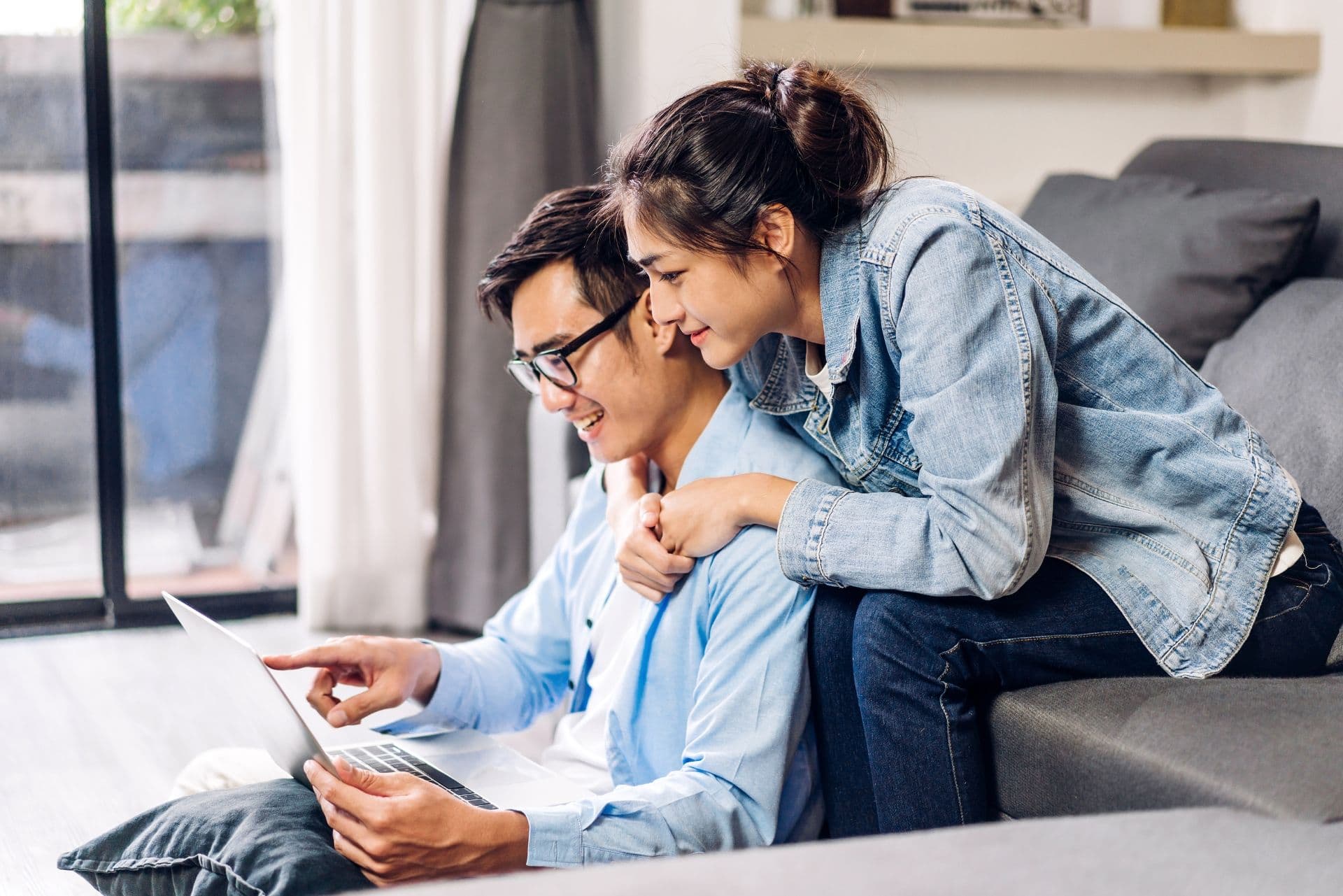 Two people sitting on a couch smiling while looking at a laptop together in a bright living room.