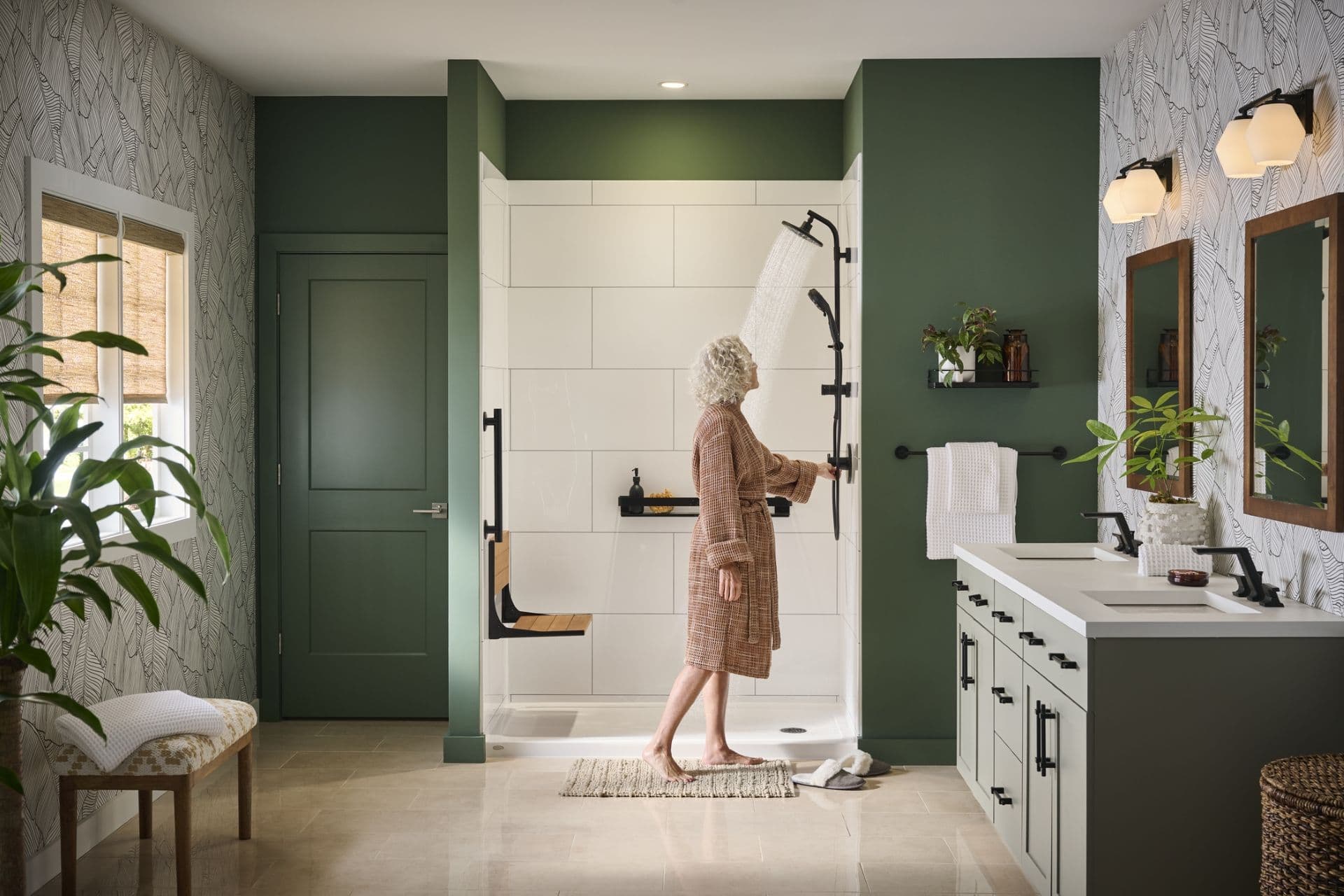 Modern bathroom with green accent walls, woman turning on a walk-in shower with white subway tile, dual vanity with black fixtures, fern-patterned wallpaper, and potted plants throughout.